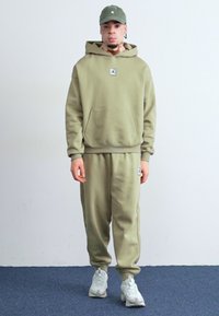 Olivengrøn hættetrøje og sweatpants sæt, lavet af blødt stof. Har en kengurulomme og logo-patch på brystet. Hvide sneakers fuldender looket.