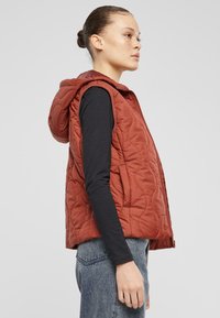 Gestepptes rotes Westchen mit Kapuze, ausgestattet mit einem Reißverschluss und seitlichen Taschen. Getragen über einem schwarzen Langarmshirt. Texturiertes Material sichtbar.