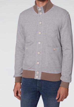 Man die een lichtgrijze vest met knopen draagt met een bruine kraag, manchetten en zoom, gecombineerd met donkerblauwe jeans, staand tegen een effen achtergrond.