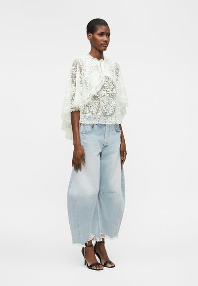 Femme portant un top en dentelle blanche transparente avec des motifs floraux, un jean large bleu clair raccourci et des sandales noires à talons hauts, debout devant un fond uni.