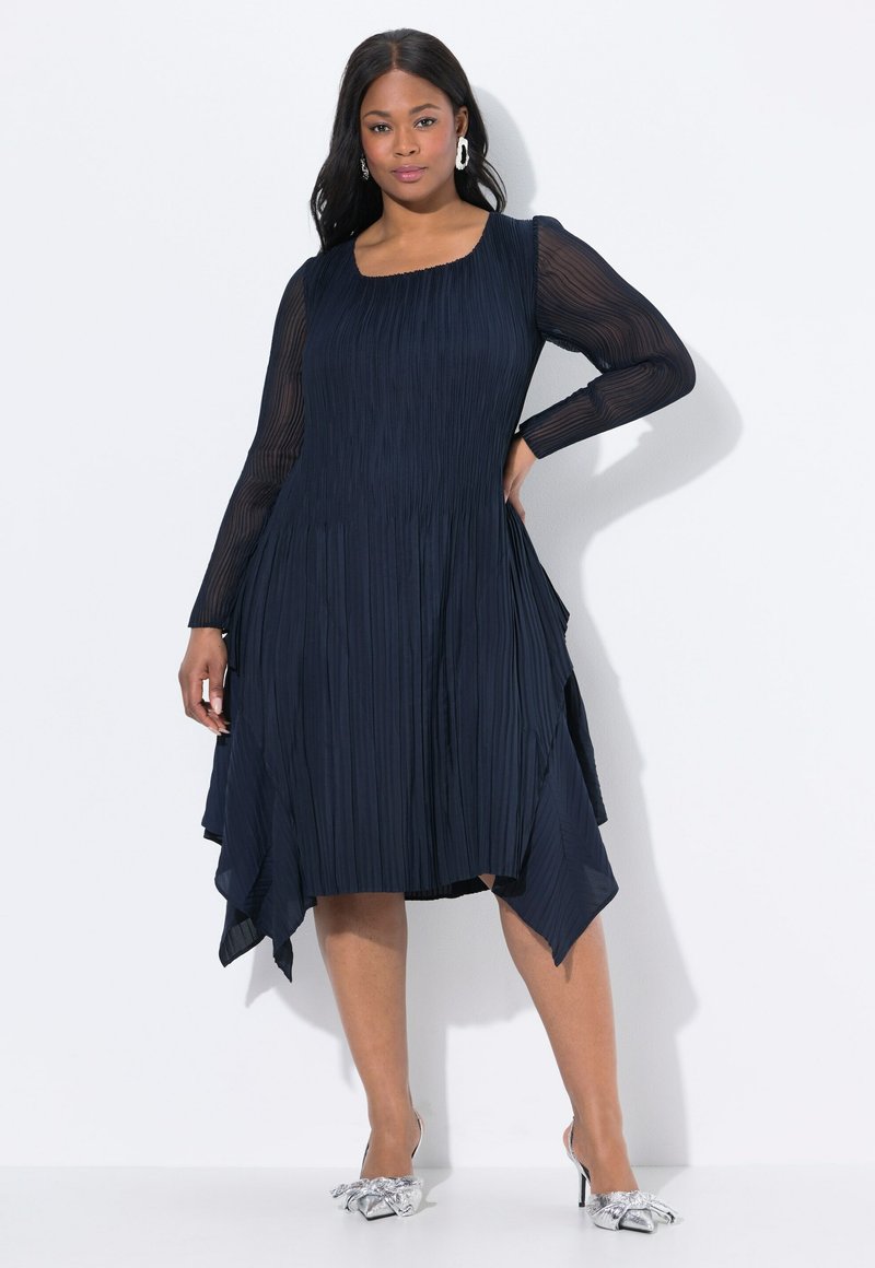 Mujer vestida con un vestido plisado azul marino de mangas largas transparentes, de pie con una mano en la cadera y tacones plateados puntiagudos con lazos.