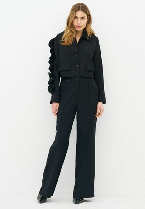 Woman with long hair wearing a black buttoned jacket with ruffles on one sleeve and matching black wide-leg pants and studded shoes.