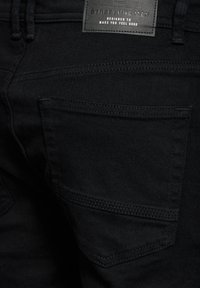 Svarta denimjeans med en texturerad yta, som har funktionella bakfickor och en etikett placerad vid midjan.