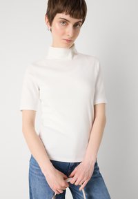Hvit t-skjorte med kort erm og høy hals laget av et glatt materiale, med glidelåsdetalj i nakken. Kombinert med blå denimjeans.