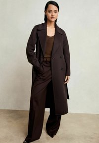 Brūns vilnas mētelis ar izgrieztu apkakli un dubultu priekšpusi, kombinēts ar tumši brūnām biksēm. Gluda tekstūra un pielāgota dizaina.