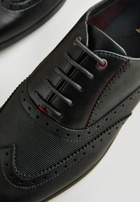 Czarne buty z syntetycznej skóry o teksturowanej powierzchni, kontrastujące czerwone szwy, sznurowany krój oraz perforowane detale na nosku.