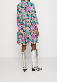 Donna che indossa un vestito a fantasia astratta colorato e stivali da cowboy metallici argento fin sopra al ginocchio, in piedi davanti a uno sfondo bianco semplice.
