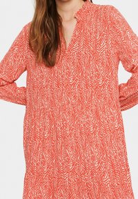 Červené šaty se vzorem, s výstřihem do V, dlouhými rukávy a nařaseným pasem. Design obsahuje bílé cik-cak pruhy na texturované látce.