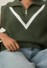 Bezrukávový svetr olivové barvy pletený s bílým V vzorem a zlatým zipem na límci, nošený přes světle modré džíny, ruka odpočívající na klíně.