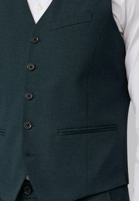 Donkergroene gebuttonte vest gedragen over een wit shirt, met vier knopen en twee horizontale zakken aan de voorkant.