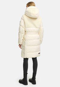 Krämfärgad pufferjacka med en plysch, strukturerad baksida, överdimensionerad huva, bälte i midjan och en svart logotypdetalj nära fållen.
