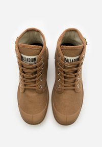 Bottines en toile marron avec bouts arrondis en caoutchouc, œillets en métal et lacets. Présente une étiquette "PALLADIUM" sur la languette. Surfaces texturées.