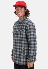 Szara i czarna flanelowa koszula w kratę z zapięciem na guziki, dwoma kieszeniami na piersi i kołnierzykiem. Noszona z czerwoną czapką. Gładka tekstura.