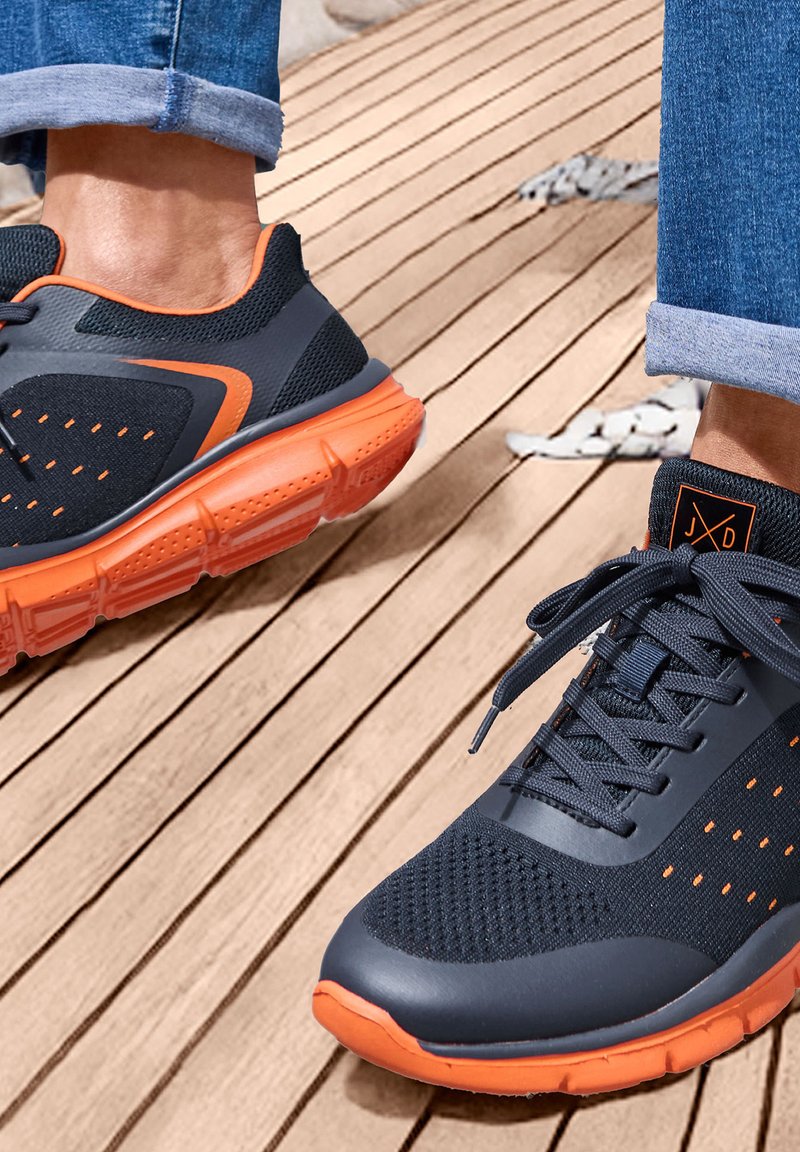 Sport-Sneaker mit einem dunklen Mesh-Obermaterial und orangefarbenen Akzenten, Gummisohle und gepolstertem Schaftrand, auf einer Holzoberfläche präsentiert.