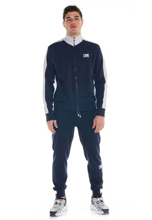 Un mannequin masculin se tient devant un fond blanc, vêtu d’un survêtement bleu marine avec des empiècements blancs et des baskets.