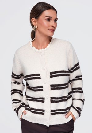 Femme portant un cardigan blanc boutonné avec des rayures horizontales noires, mains dans les poches, et des boucles d’oreilles dorées en forme de feuille, regardant sur le côté.