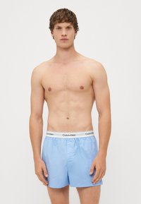 Svetlo modri bokseric z elastičnim pasom, ki ima bel logotip "Calvin Klein". Gladek material, preprost dizajn, dolžina do kolen.