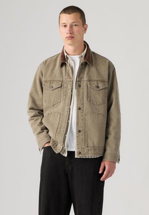 Junger Mann trägt eine khakifarbene Jacke mit Klapptaschen über einem weißen Hemd und schwarzen Hosen, steht vor einem schlichten hellen Hintergrund.