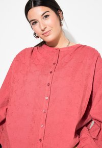 Camicia coral a vestibilità larga con scollatura rotonda, chiusure con bottoni e un motivo di ricamo floreale texturizzato su tutta la superficie.