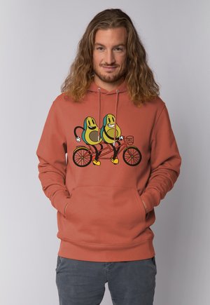 Mann mit langen Haaren, der einen rostfarbenen Hoodie trägt, auf dem zwei lächelnde Avocados abgebildet sind, die zusammen auf einem Tandemfahrrad fahren.