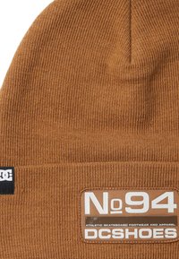 Bonnet en tricot marron avec une texture côtelée, arborant un patch cousu portant l'inscription "No 94 DC Shoes" en texte blanc sur un fond beige.
