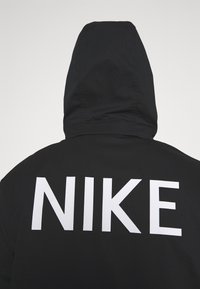 Black hoodie with adjustable hood, featuring a large white "NIKE" logo across the back. Material appears lightweight and textured.