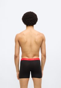 Μαύρα αντρικά εσώρουχα boxer με κόκκινη μέση που εμφανίζει το μήνυμα "TOMMY HILFIGER" σε λευκό κείμενο. Λείο ύφασμα, εφαρμοστή σχεδίαση. Το μοντέλο φαίνεται από πίσω.