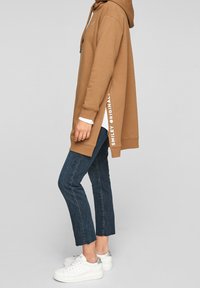 Sudadera oversize marrón con aberturas laterales, que presenta el texto blanco "SMILEY ORIGINAL". Combinada con jeans de lavado oscuro ligeramente recortados y zapatillas blancas.