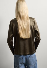 Satin-olivgrüne Jacke mit langen Ärmeln, rechteckiger Form, glatter Textur und runder Saumnaht, kombiniert mit hellblauen Denim-Jeans.