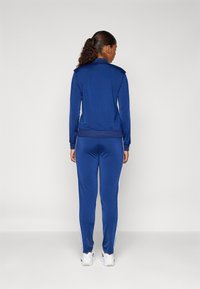 Blå idrottsoverall tillverkad av slätt tyg. Har en zip-jacket, byxor med elastisk midja och vita sidosträck. Ren design utan mönster.