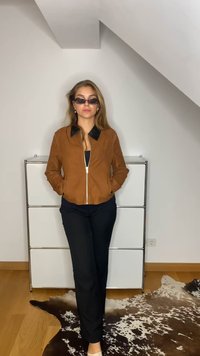 Brown suede jacket with a black collar, black pants, and sunglasses. Standing against a neutral wall, with a white dresser in the background.