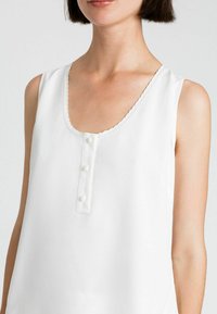 Top bianco senza maniche con scollatura arrotondata, rifinitura decorativa a coste e tre bottoni sul davanti. Stoffa liscia con una leggera lucentezza.