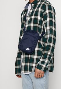 Námořnická nylonová crossbody taška s nastavitelnými popruhy a bílým logem Nike. Kompaktní obdélníkový tvar, vybavená zapínací kapsou a měkkou texturou.