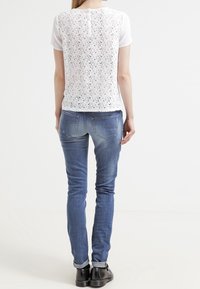 Top bianco a maniche corte con retro in pizzo, abbinato a jeans in denim blu aderenti e scarpe nere. Design semplice, senza motivi visibili.
