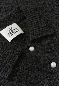 Črno teksturirano blago z rebrastim ovratnikom, z dvema belima bisernima gumboma in oznako s črnim logotipom na levi strani.