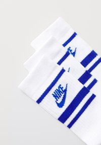 Chaussettes de sport blanches avec des rayures bleues et un logo brodé. Présente des bords côtelés et un tissu doux et texturé. Trois paires visibles.