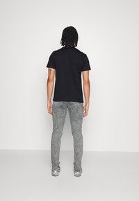 Camiseta negra de manga corta, jeans skinny lavados en gris y zapatillas plateadas; sujeto de espaldas con cabello corto y texturizado y un tatuaje en el brazo.