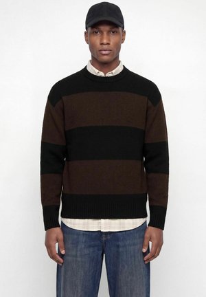 Homme portant un pull rayé noir et brun foncé, une chemise à carreaux claire en dessous, un jean bleu et une casquette noire, debout devant un fond blanc.