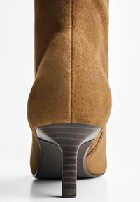Ruske nubuk ankle boot s visokom potpeticom. Ima glatku teksturu i vertikalne šavove na poleđini. Minimalistički dizajn.