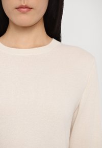 Maglione beige chiaro con un colletto rotondo a coste, maniche lunghe e una texture morbida. Vestibilità aderente con design minimalista.