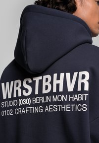 Hoodie bleu marine en tissu texturé avec un grand imprimé graphique blanc au dos, comportant "WRSTBHVR" et d'autres détails.