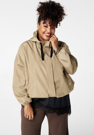 Lachende vrouw met krullend haar, een beige jack, een zwart topje met kanten rand, een bruine broek en armbanden, poserend tegen een witte achtergrond.