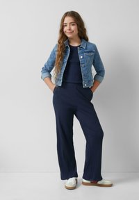 Giacca di jeans in blu chiaro con dettagli a bottoni, abbinata a un tuta a coste blu scuro e sneakers bianche con accenti marroni.