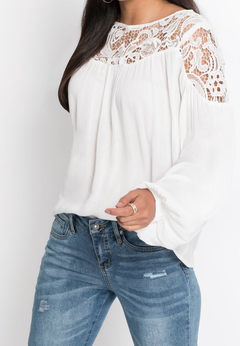 Femme portant un chemisier blanc avec des détails en dentelle aux épaules et à l’encolure, assorti d’un jean bleu et de bagues dorées à la main.