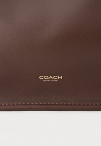 Smeđa kožna novčanik s glatkom teksturom, s reljefnim logotipom "COACH NEW YORK" u zlatu. Vidljivi šavovi.