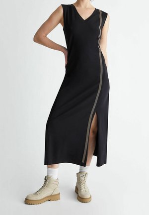 Femme portant une robe midi noire sans manches avec une fente latérale et un détail de rayures diagonales, associée à des chaussettes blanches et des bottines beiges à lacets.