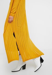 Dlouhý žebrovaný kardigan v jasně žluté barvě s kovovými nitěmi. Otevřený přední díl s bočními rozparky. Kombinován s bílými kotníkovými botami na podpatku.