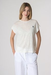 Top corto bianco leggero e semitrasparente con maniche corte e scollo a girocollo, dotato di un piccolo logo sul lato sinistro del petto. Abbinato a pantaloni bianchi larghi.