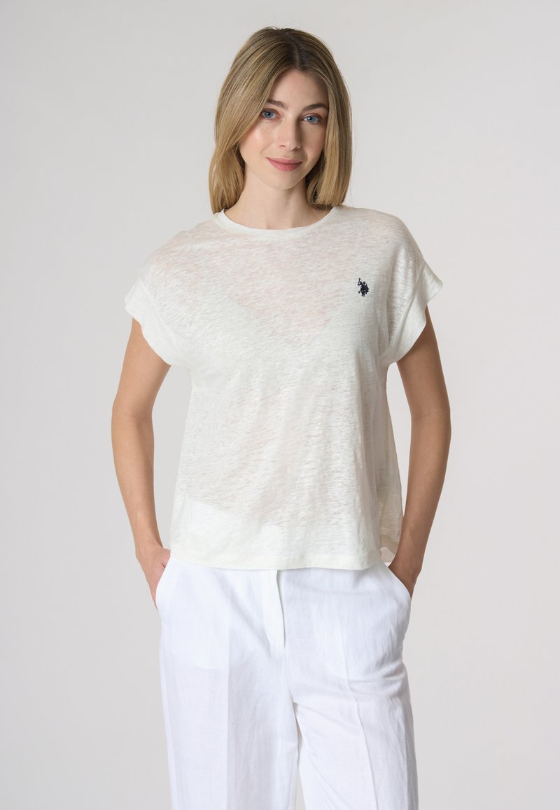 Top corto bianco leggero e semitrasparente con maniche corte e scollo a girocollo, dotato di un piccolo logo sul lato sinistro del petto. Abbinato a pantaloni bianchi larghi.