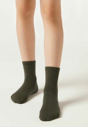 Die unteren Beine eines Kindes in dunkelgrünen Knöchelsocken, die auf einer schlichten hellen Oberfläche stehen.